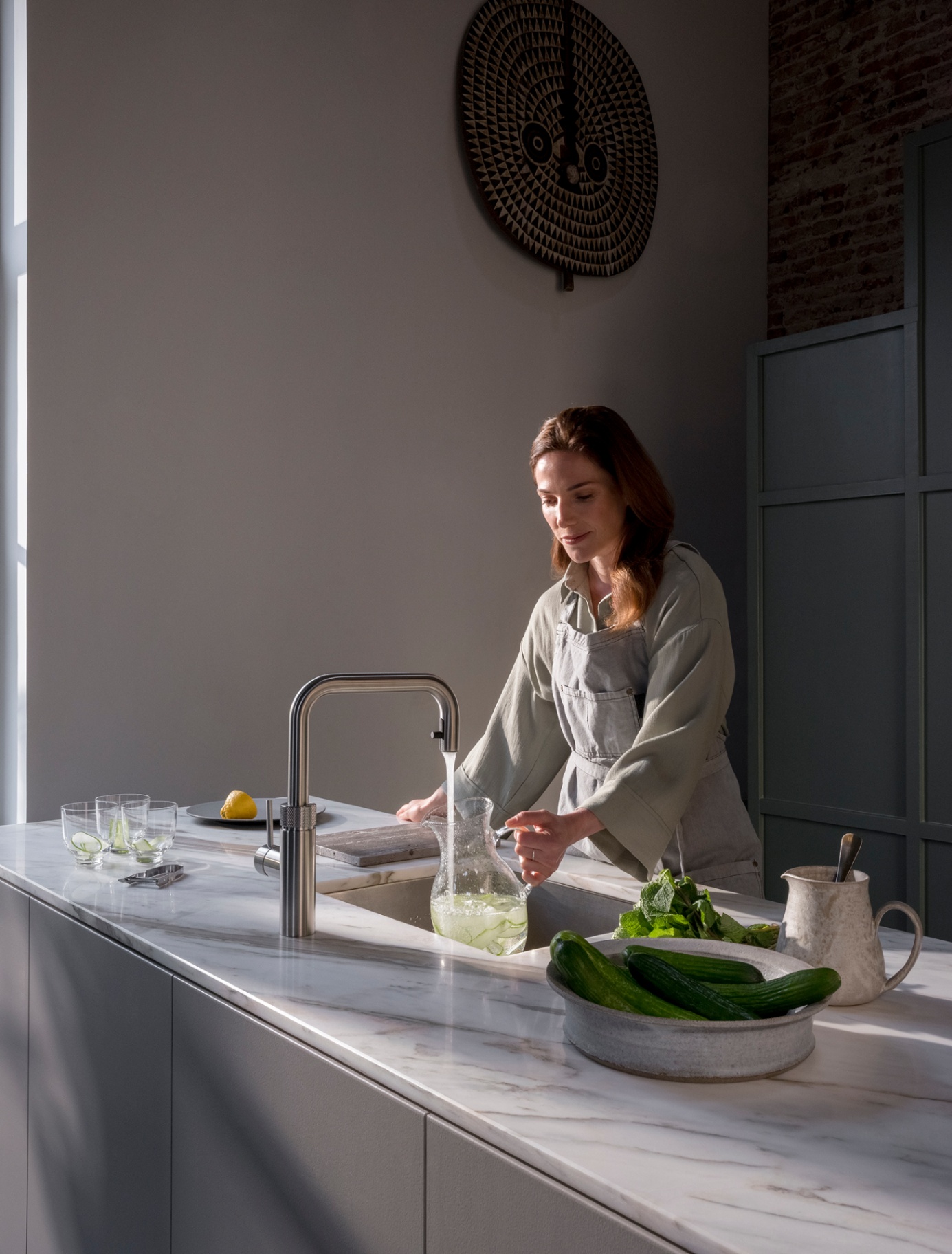 Woman filling a cucumber-infused water carafe at a Flex Square stainless steel Quooker on marble