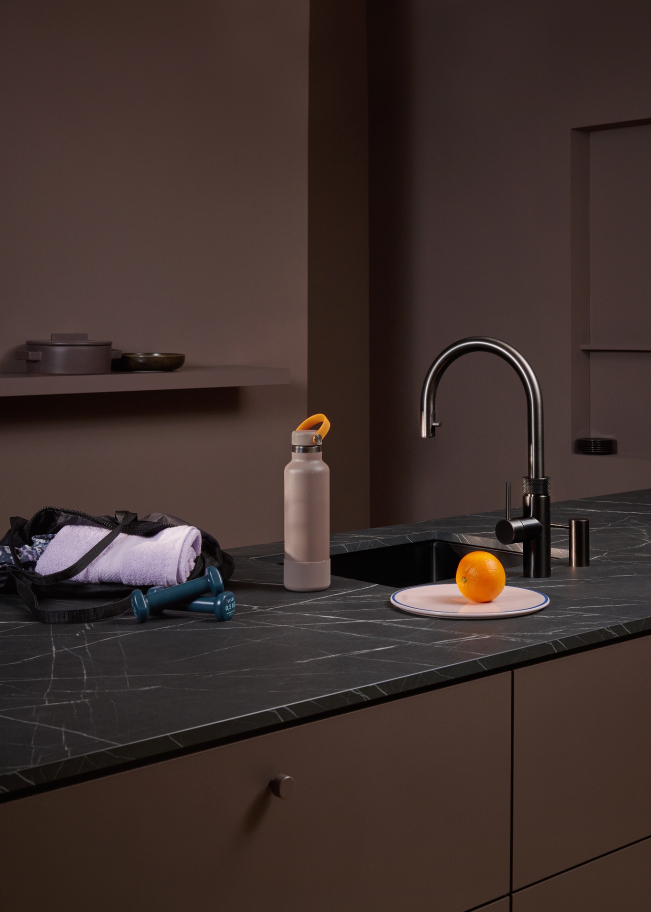 Still life of a Flex Round gun metal Quooker with a sports bottle and orange on dark marble
