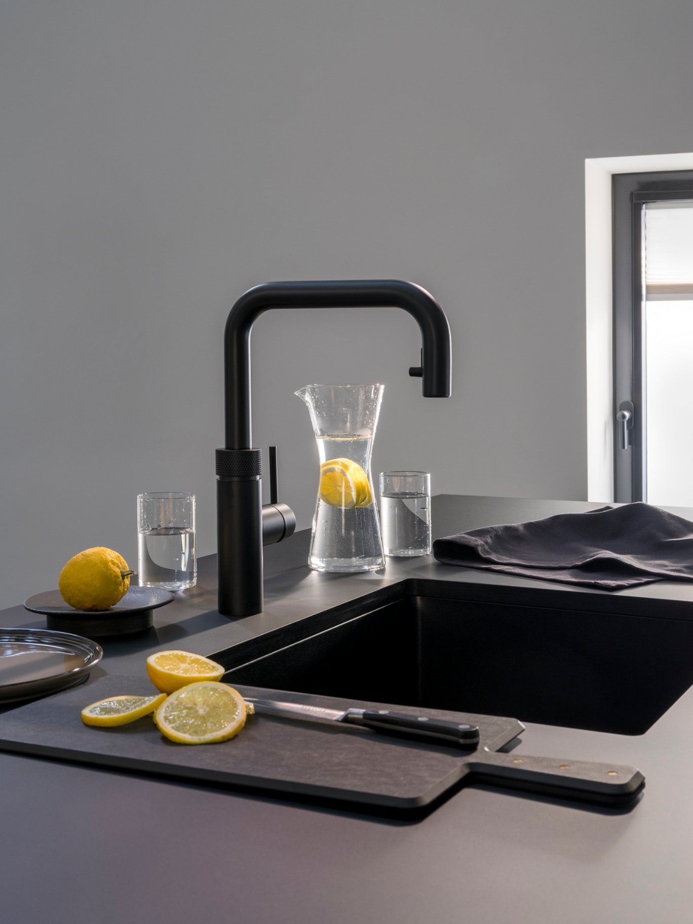 Still life of a Flex Square black Quooker with lemon-infused water carafe and sliced lemons on a matte black worktop