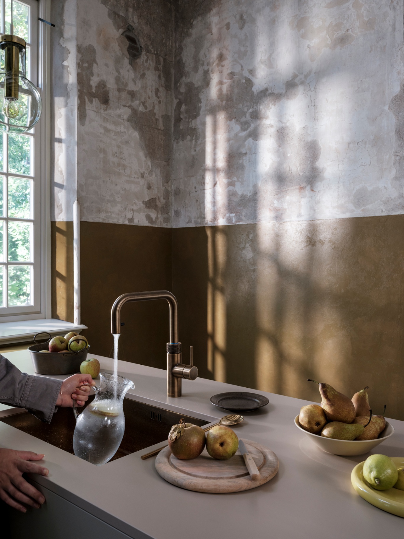 Woman filling a glass carafe with sparkling water at a Flex Square patinated brass Quooker in a moody atelier