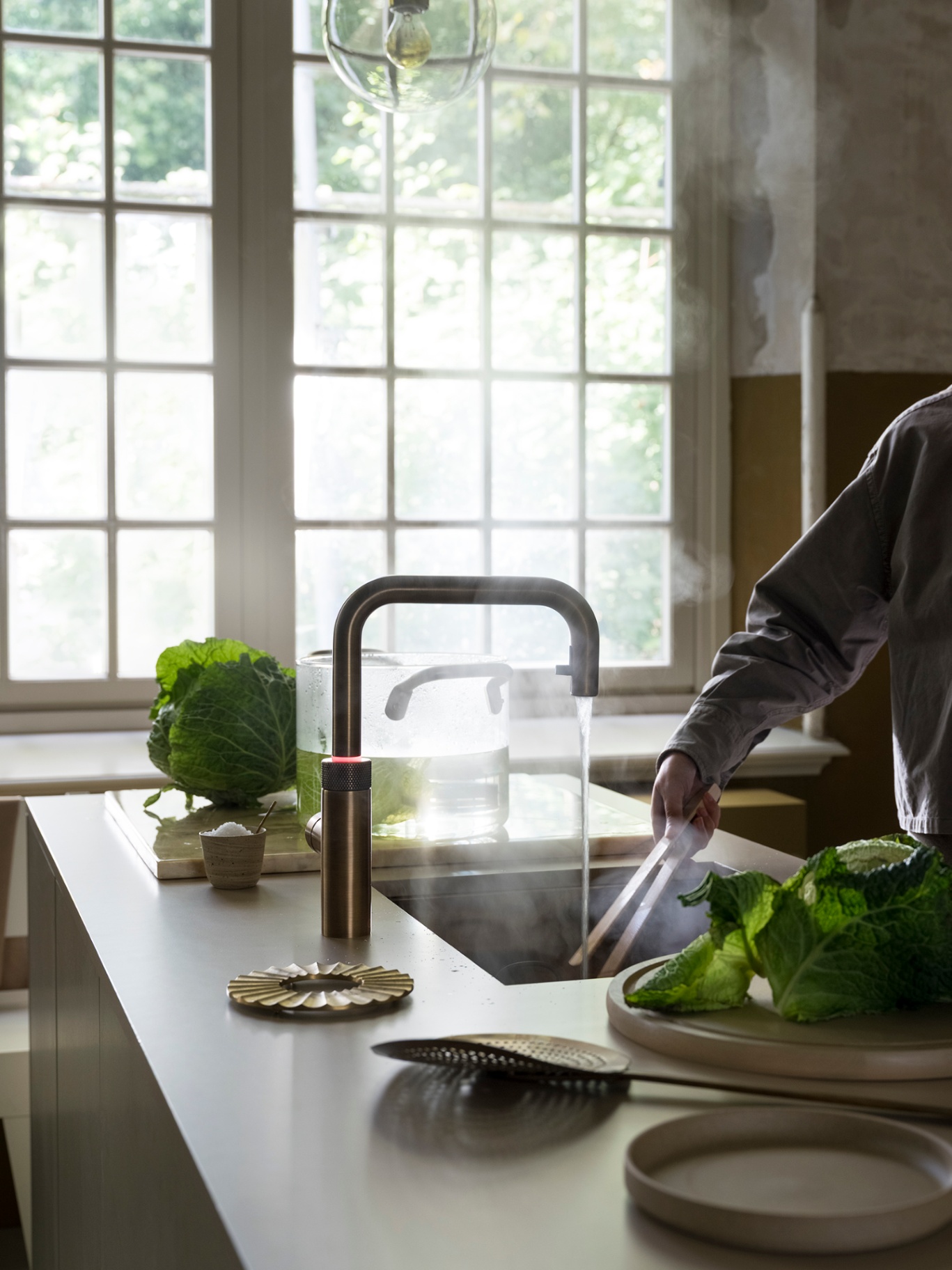 Wide editorial view of a Flex Square patinated brass Quooker steaming above a glass pot with savoy cabbage