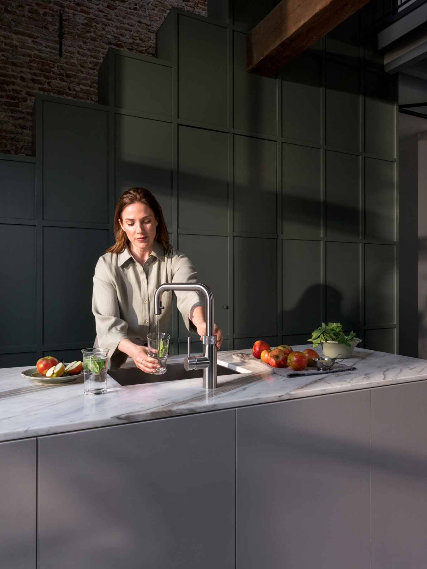 Woman filling a glass of chilled mint water at a Flex Square stainless steel Quooker with apples on marble