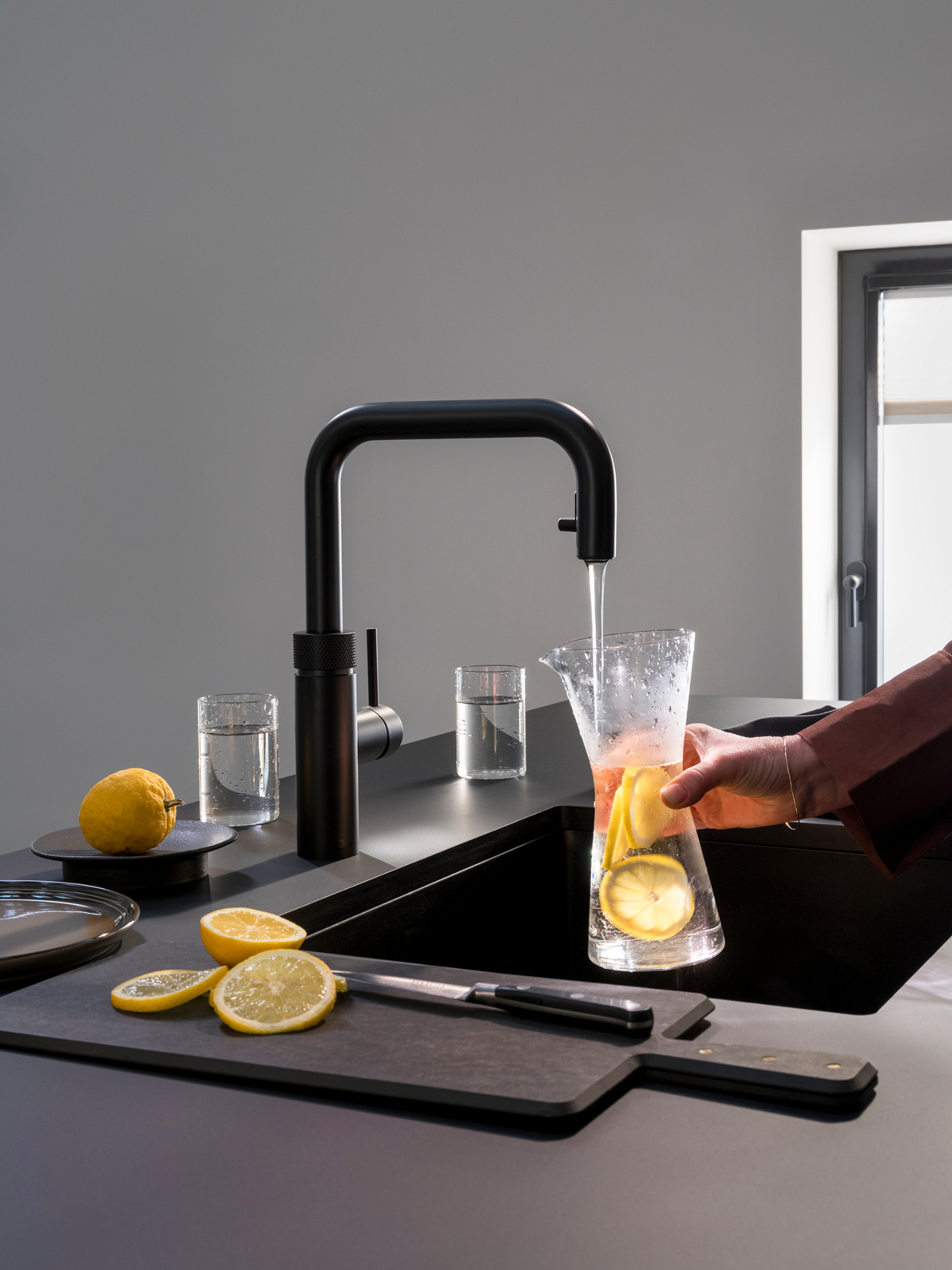 Hand pouring chilled lemon water from a Flex Square black Quooker on a dark island