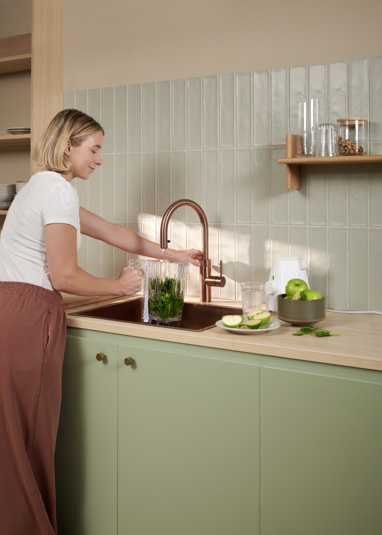 Woman preparing a green smoothie pitcher under a Flex Round rose copper Quooker in a sage tile kitchen