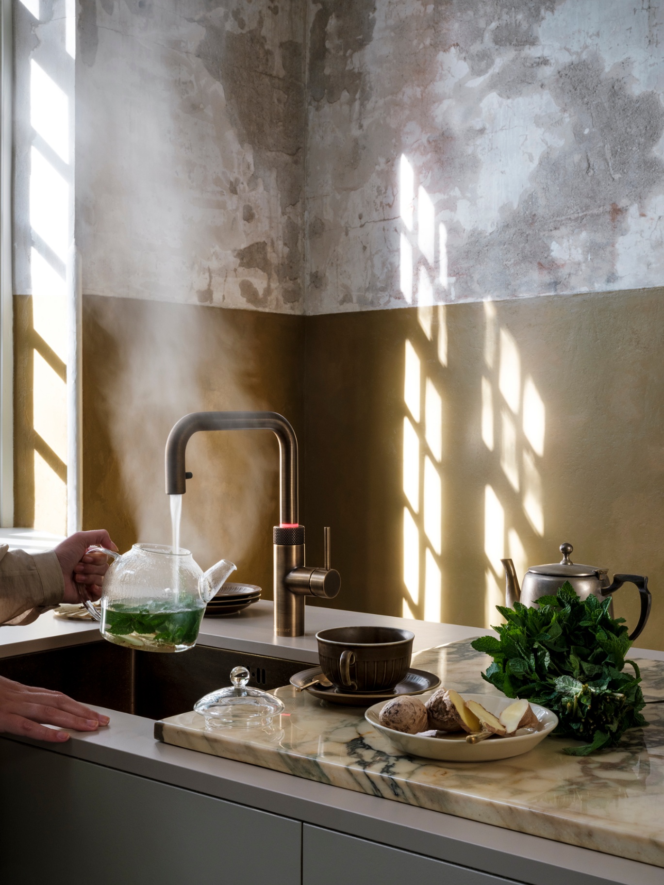 Woman brewing fresh mint tea in a glass teapot at a Flex Square patinated brass Quooker with red LED ring