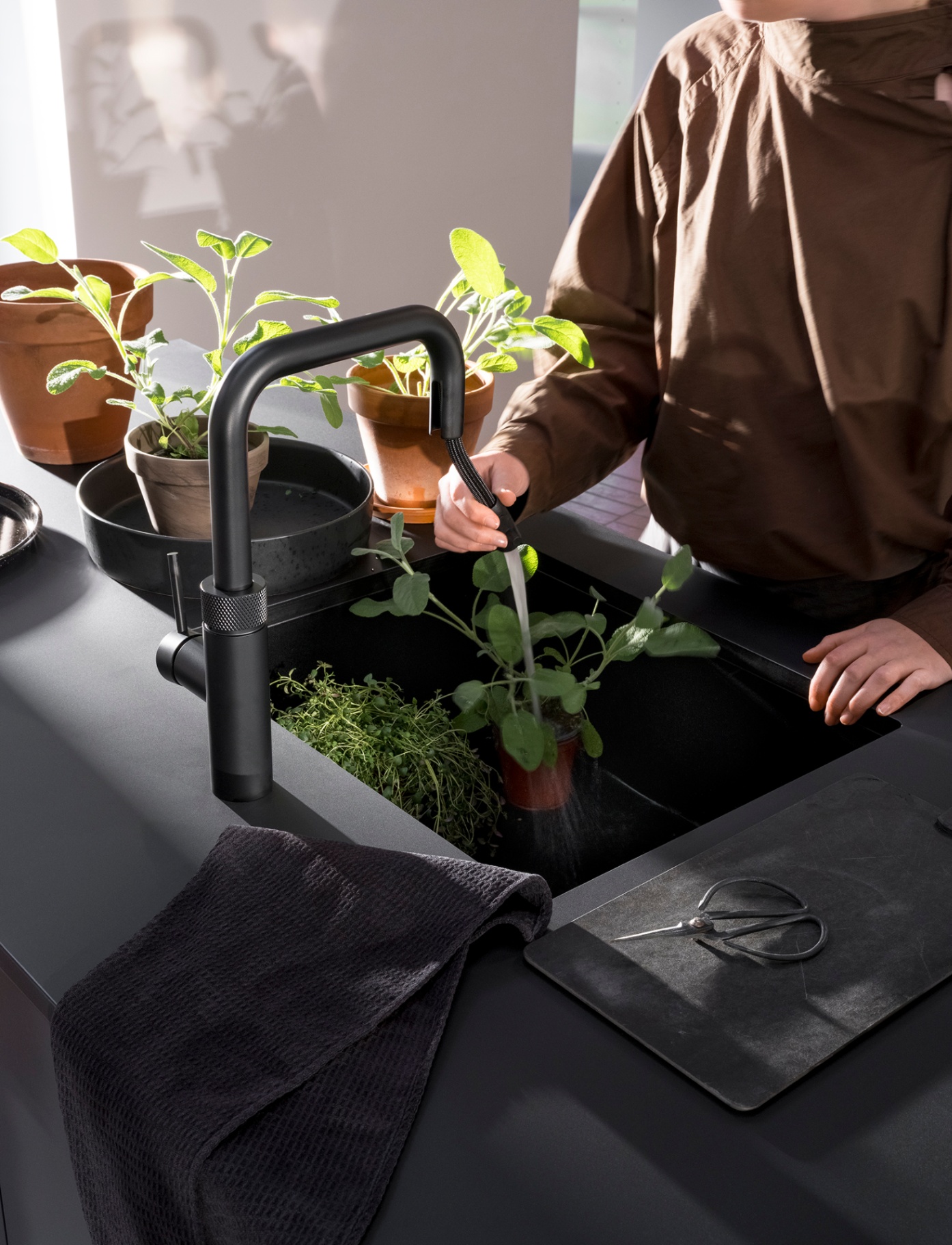 Woman rinsing potted herbs with the pull-out hose of a Flex Square black Quooker on a matte black kitchen island