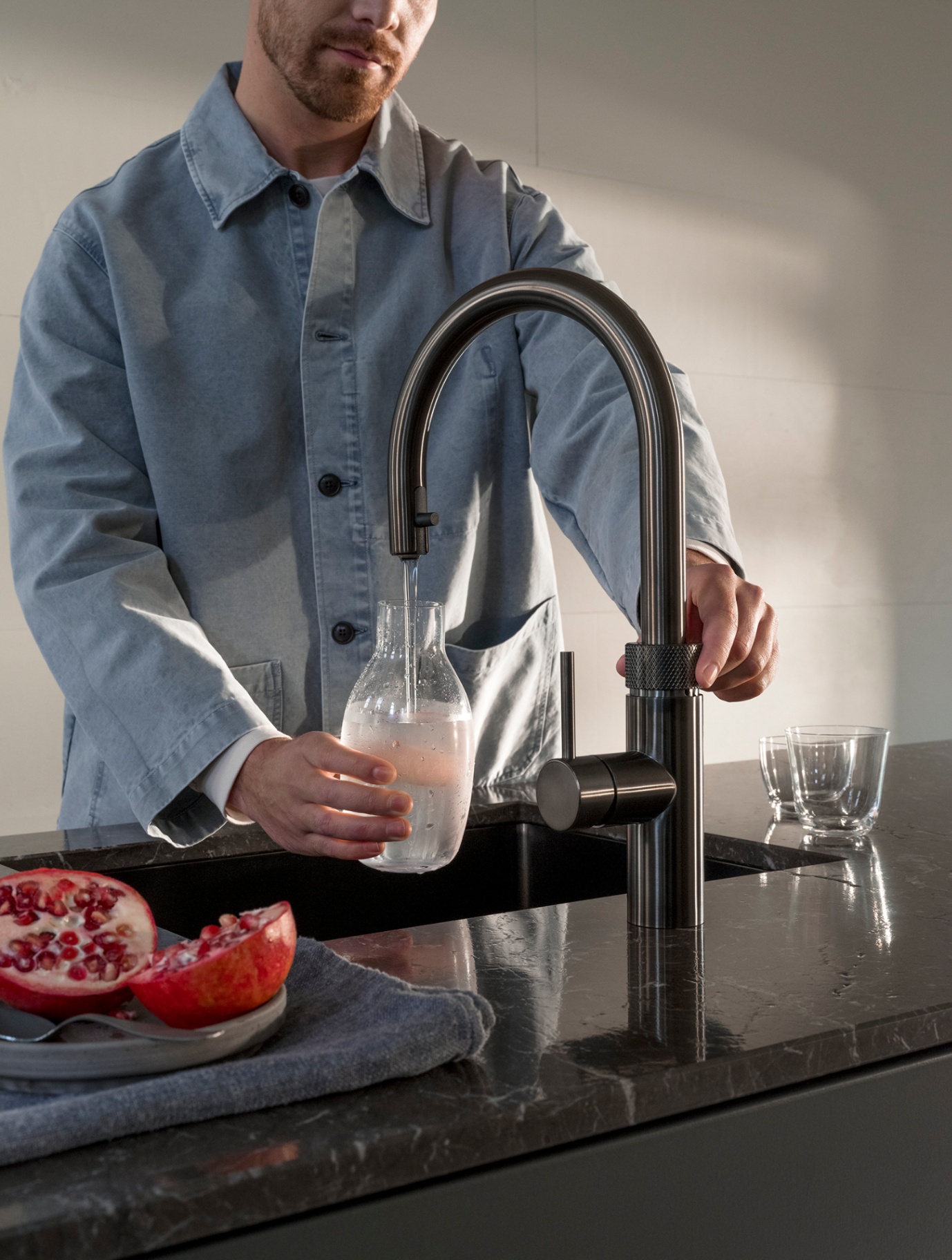 Man filling a carafe of chilled water at a Flex Round gun metal Quooker on black marble beside pomegranate