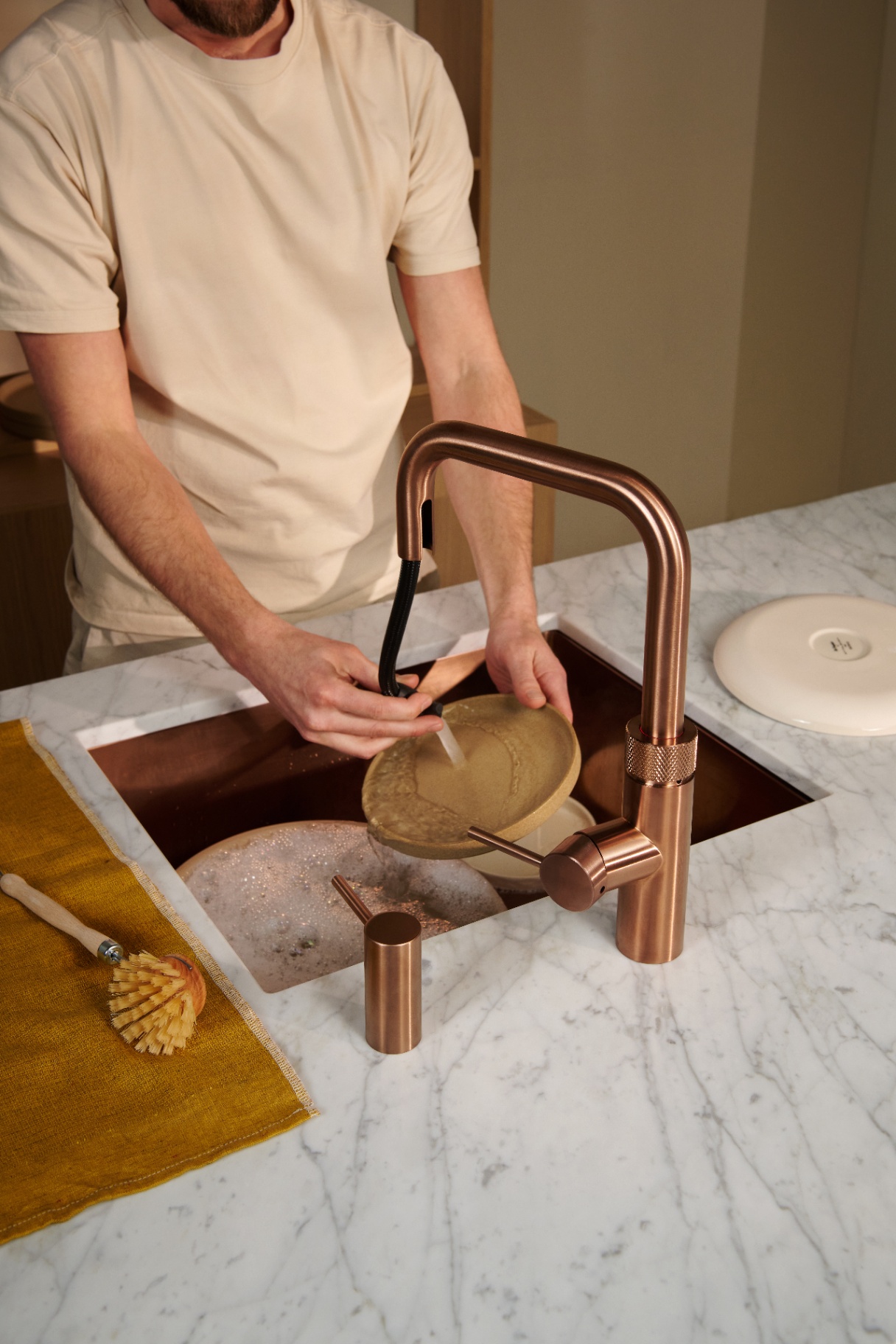 Man rinsing a ceramic plate with the pull-out hose of a Flex Square rose copper Quooker on Carrara marble