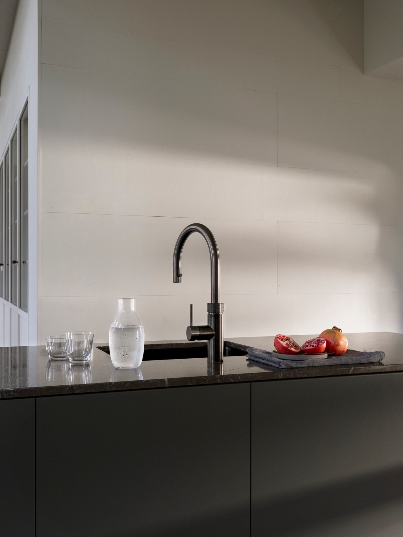 Editorial still of a Flex Round gun metal Quooker on dark marble with a carafe of still water and pomegranate
