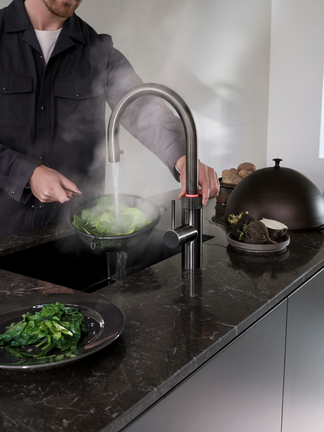 Close-up of a man boiling spinach at a Flex Round gunmetal Quooker with red LED safety ring on dark marble