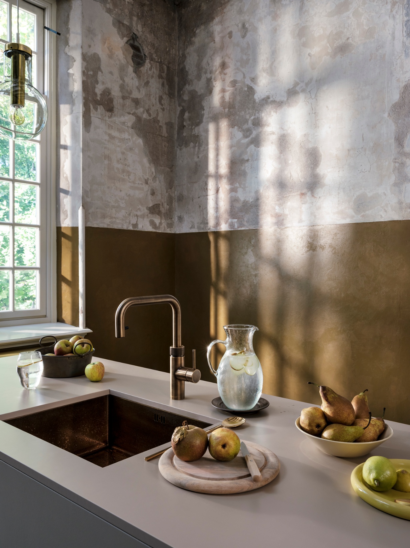 Editorial still of a Flex Square patinated brass Quooker with a sparkling water carafe, pears and apples on stone