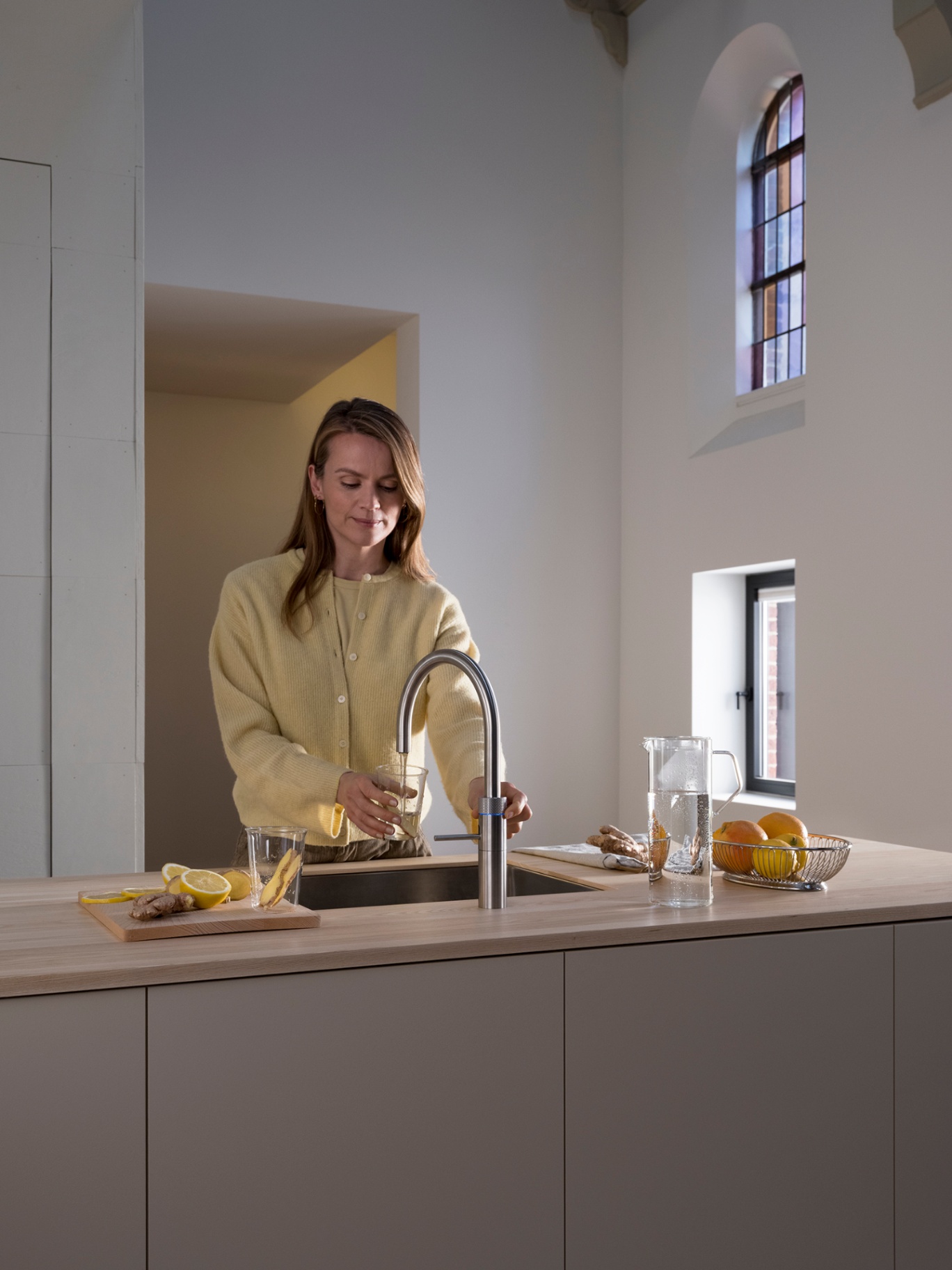 Woman filling a glass with chilled lemon-ginger water at a Fusion Round stainless steel Quooker in a cathedral-like kitchen