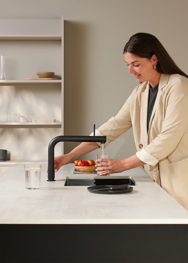 Quooker Front in matte black boiling water tap — pouring filtered sparkling water, with fresh pear in a real customer kitchen. Genuine Quooker product supplied by Watertap.eu.