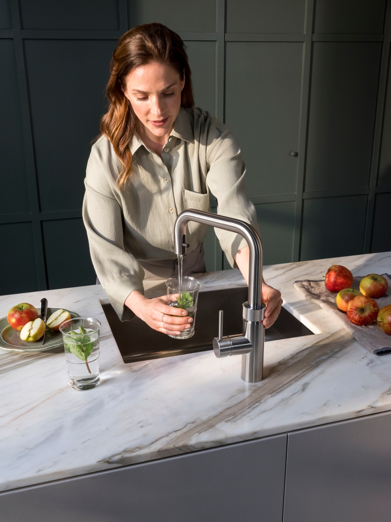 Overhead of a woman filling a glass of chilled mint water at a Flex Square stainless steel Quooker with red apples