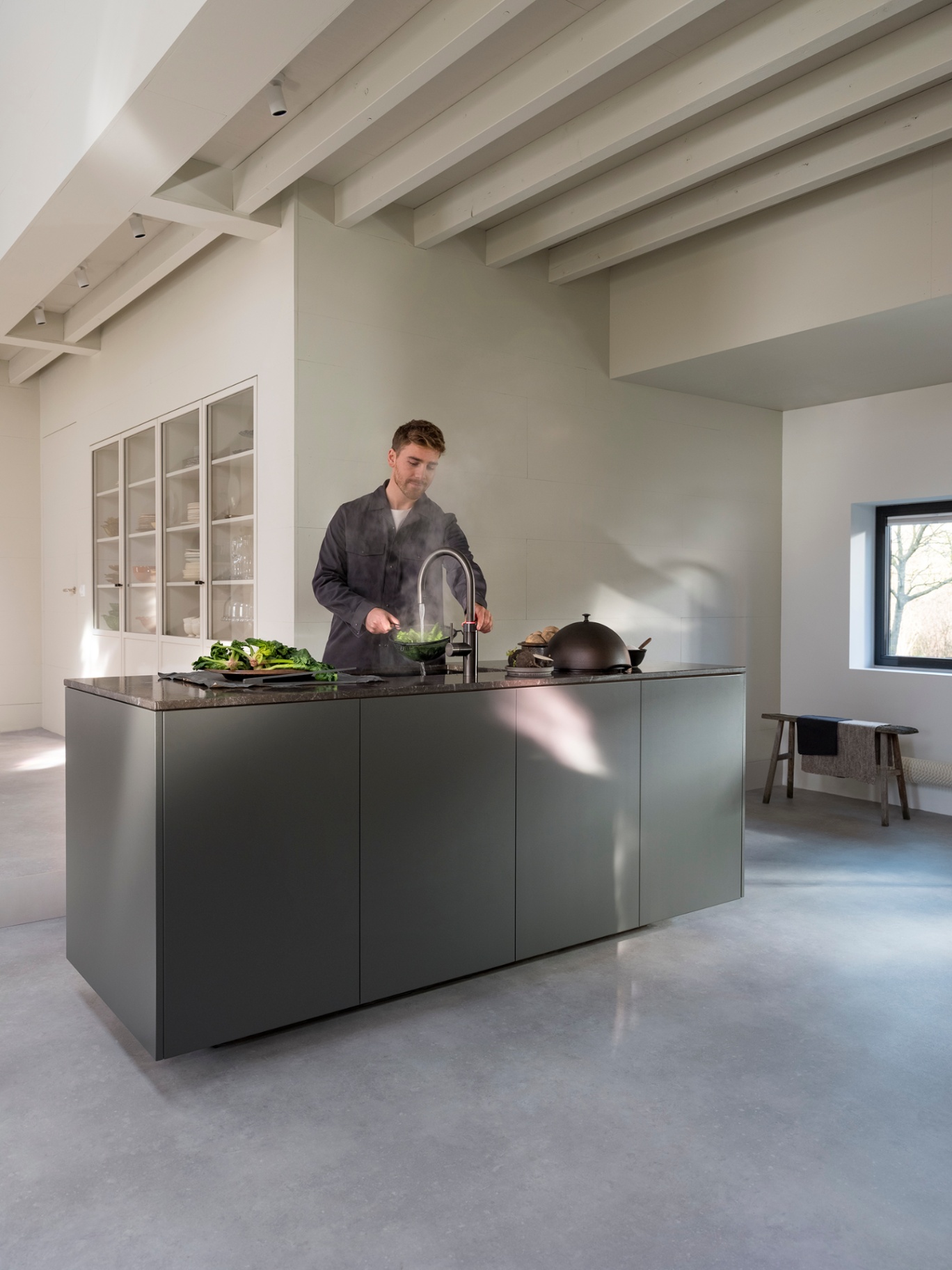 Man boiling leafy greens at a Flex Round gun metal Quooker on a dark marble island in a bright open kitchen
