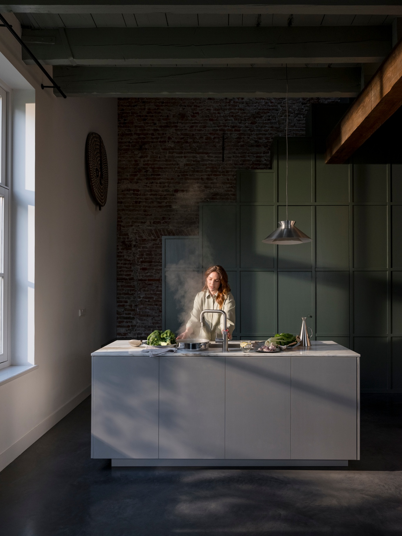 Wide loft kitchen shot of a woman cooking greens at a Flex Square stainless steel Quooker with steam rising