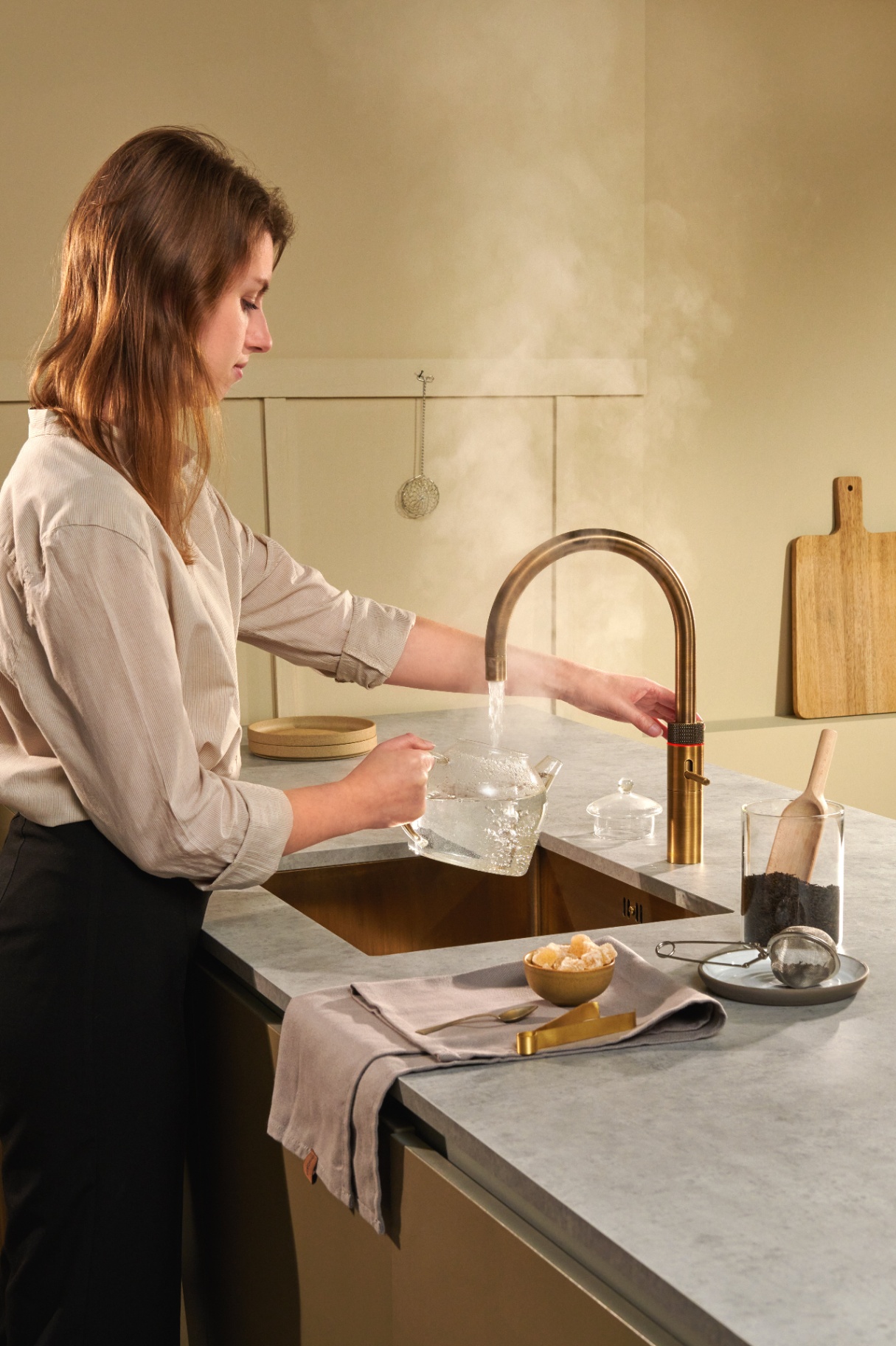 Woman brewing loose-leaf tea below a Fusion Round patinated brass Quooker in a warm ochre kitchen